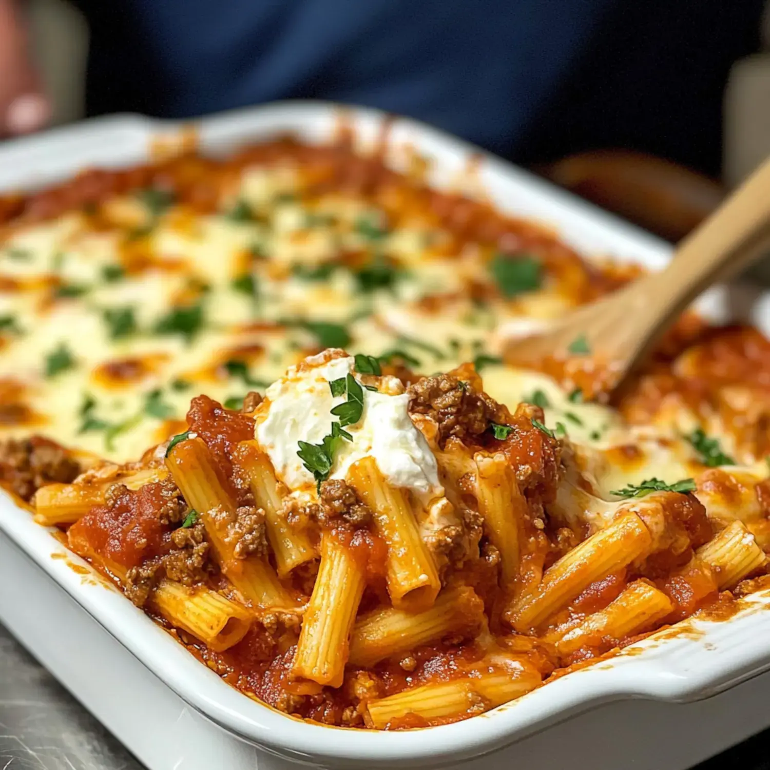 A close-up of a serving of baked pasta with marinara sauce, ground meat, and topped with cheese and herbs in a white dish.