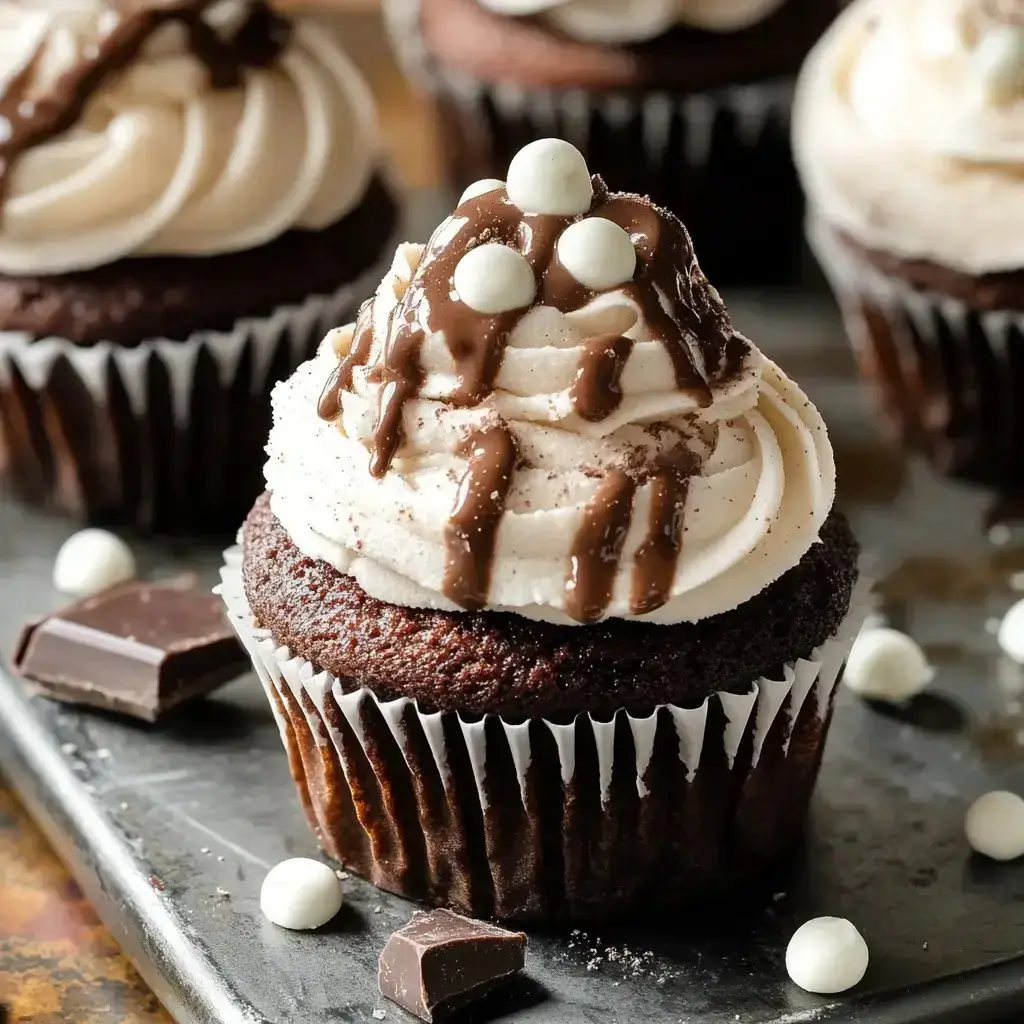 A close-up of a chocolate cupcake topped with swirls of cream frosting, drizzled chocolate, and white chocolate candies, surrounded by additional cupcakes and chocolate pieces on a dark surface.