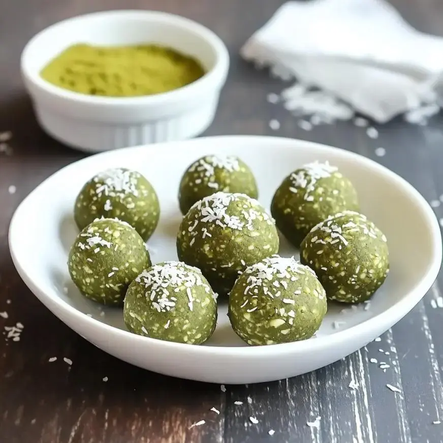 A white bowl filled with green energy balls sprinkled with shredded coconut, with a small dish of green powder in the background.