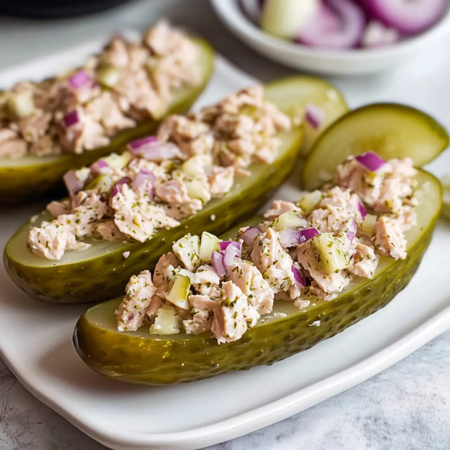 Three sliced pickles filled with a mixture of shredded chicken, diced onions, and herbs are displayed on a white plate.