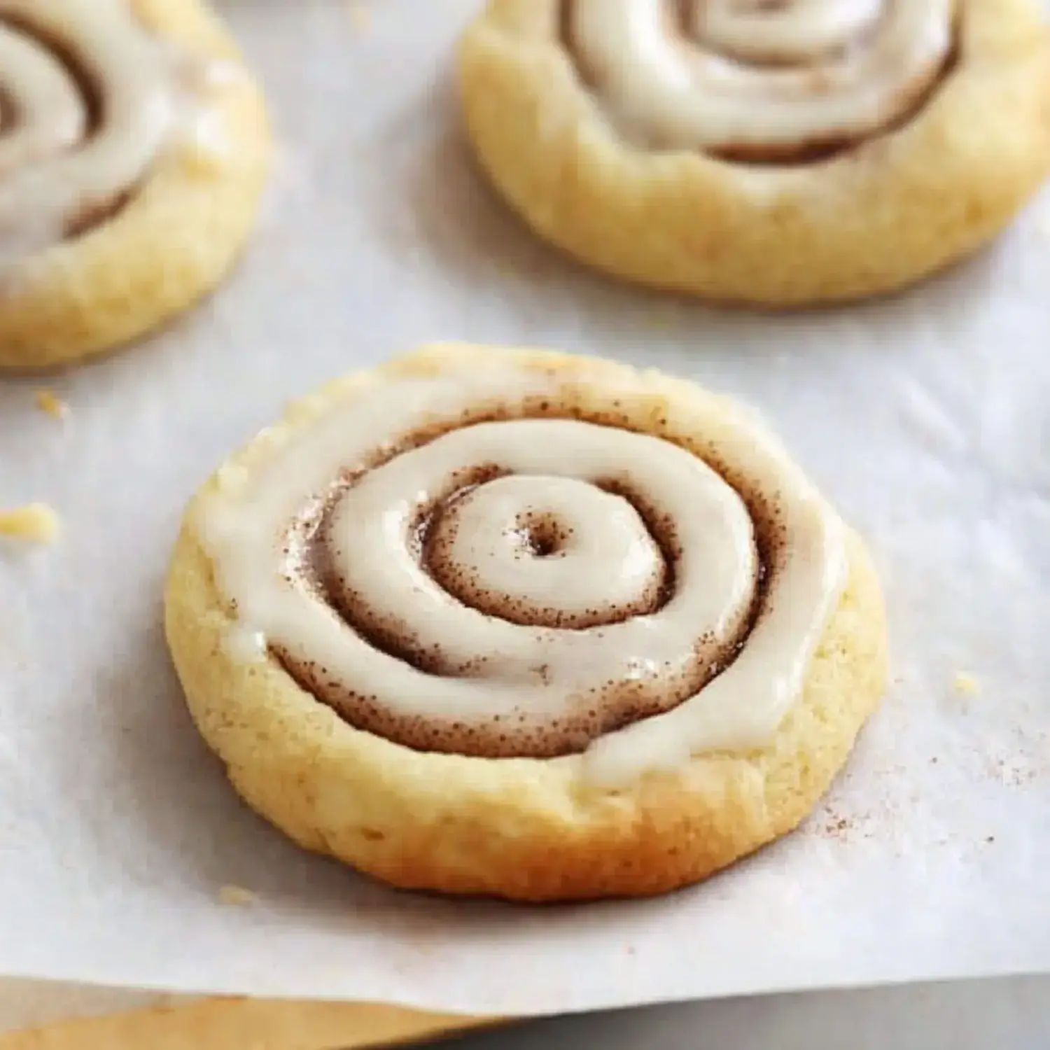 A close-up image of a cinnamon roll cookie topped with creamy icing, sitting on parchment paper.