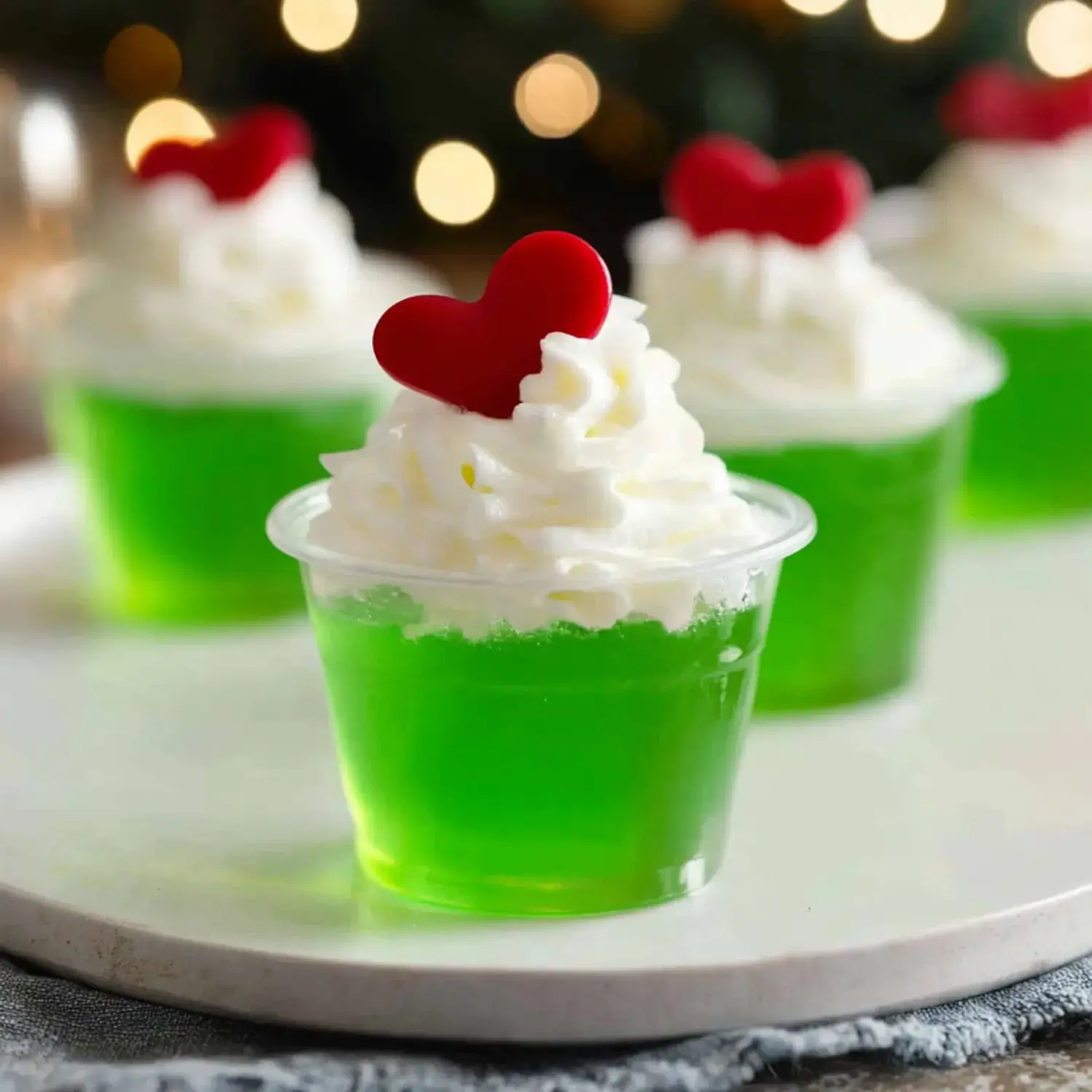 A close-up of green gelatin desserts topped with whipped cream and red heart-shaped decorations, set against a blurred background of festive lights.