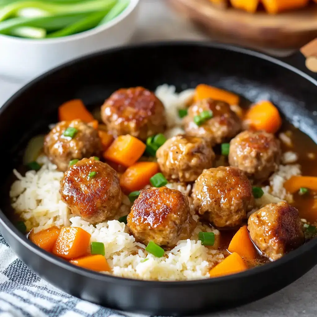 A black skillet contains cooked meatballs, diced carrots, green onions, and white rice all coated in sauce.