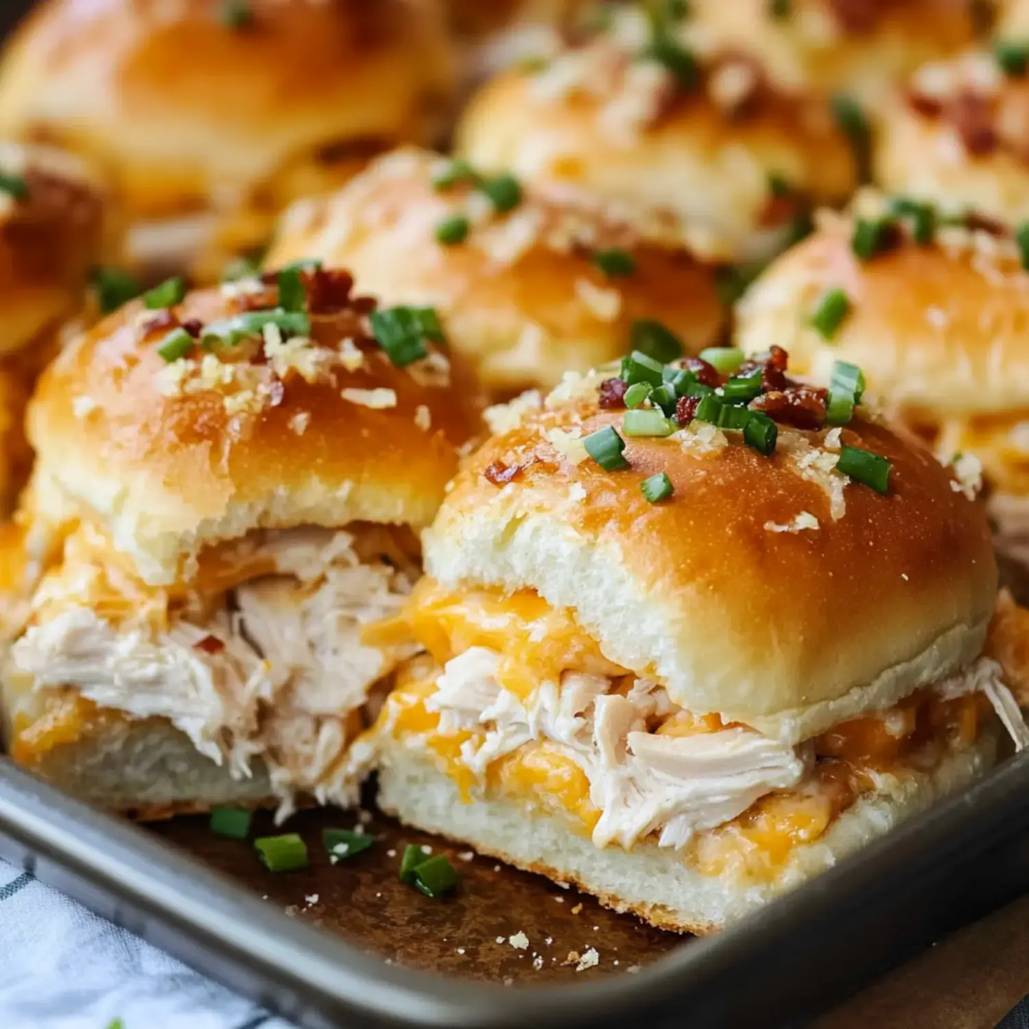 A close-up of fluffy slider sandwiches filled with shredded chicken and melted cheese, topped with green onions and crispy bits, arranged on a tray.