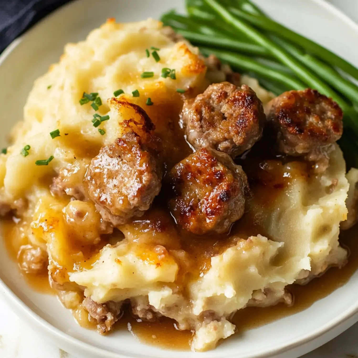 A plate of creamy mashed potatoes topped with savory meatballs and gravy, alongside fresh green beans.