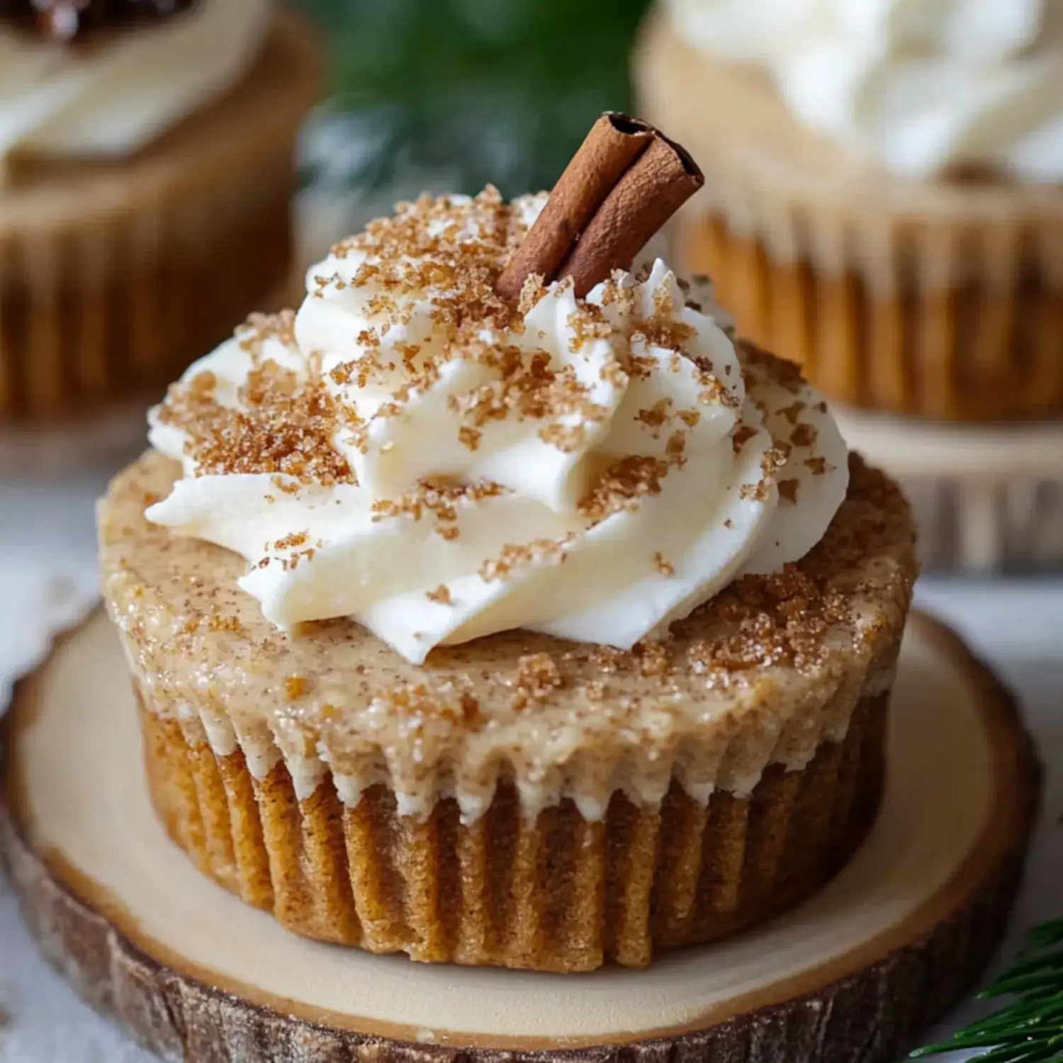 A close-up of a cinnamon-flavored cupcake topped with whipped cream, brown sugar, and a cinnamon stick, set on a wooden slice.