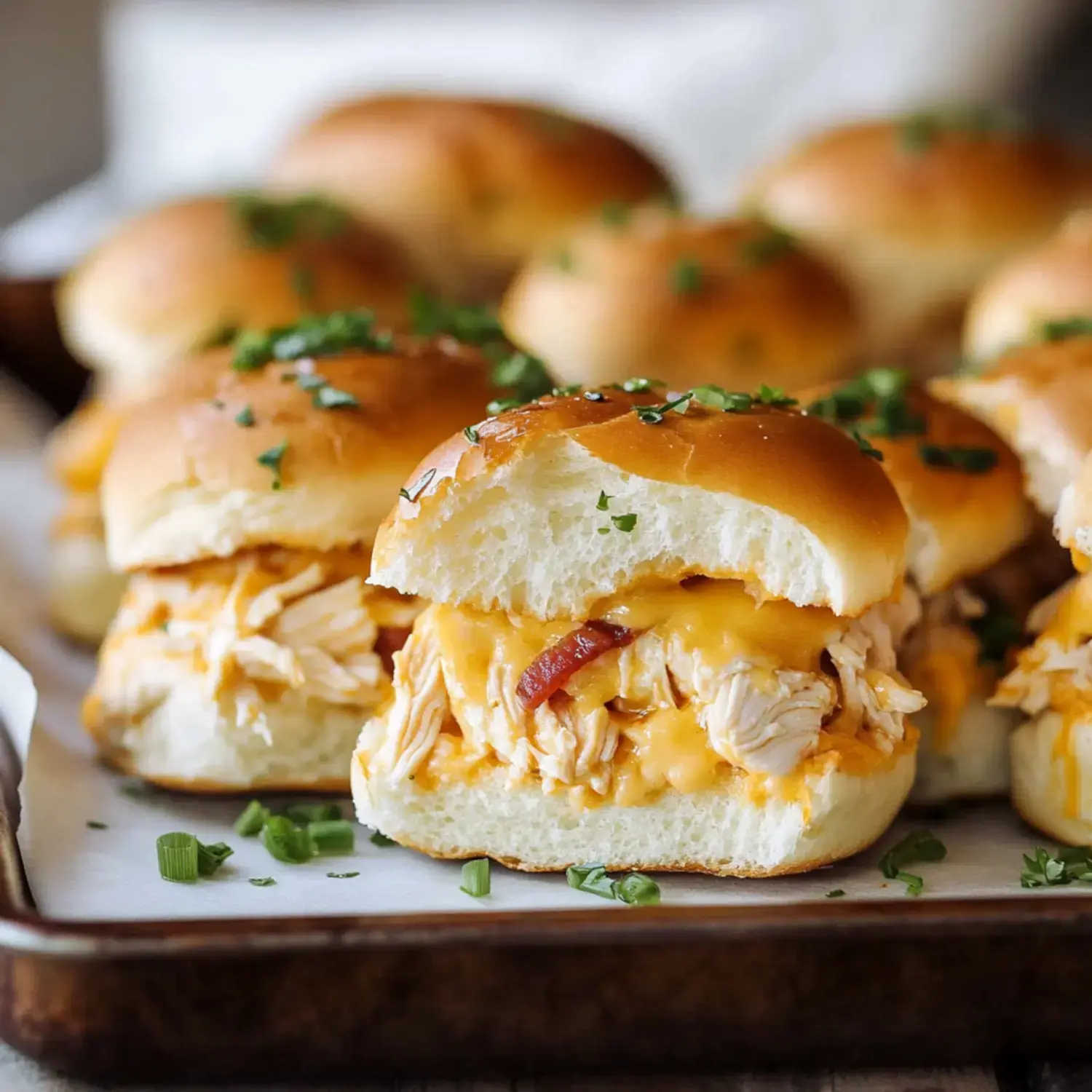 A close-up of a pulled chicken sandwich with melted cheese and a bite taken out, surrounded by additional sandwiches garnished with green herbs.