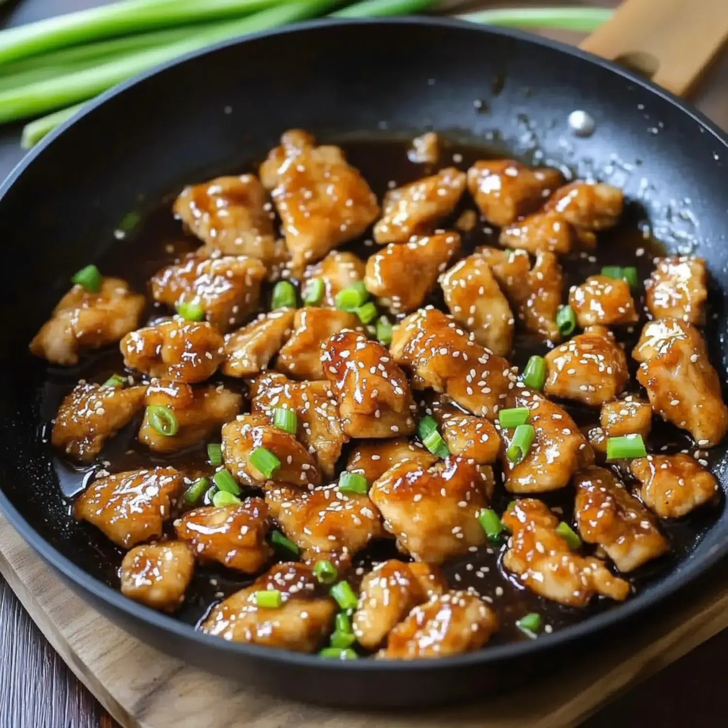 A skillet filled with glazed chicken pieces garnished with green onions and sesame seeds.