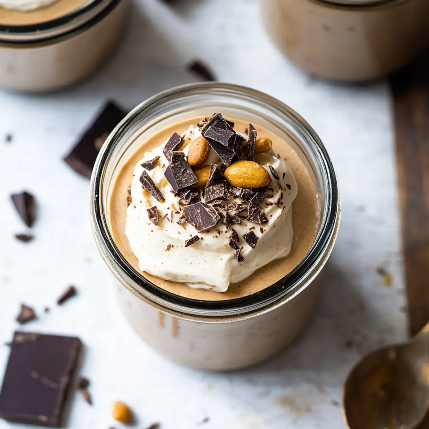 A close-up of a glass jar filled with creamy dessert topped with whipped cream, chopped chocolate, and almonds, with pieces of chocolate scattered around.