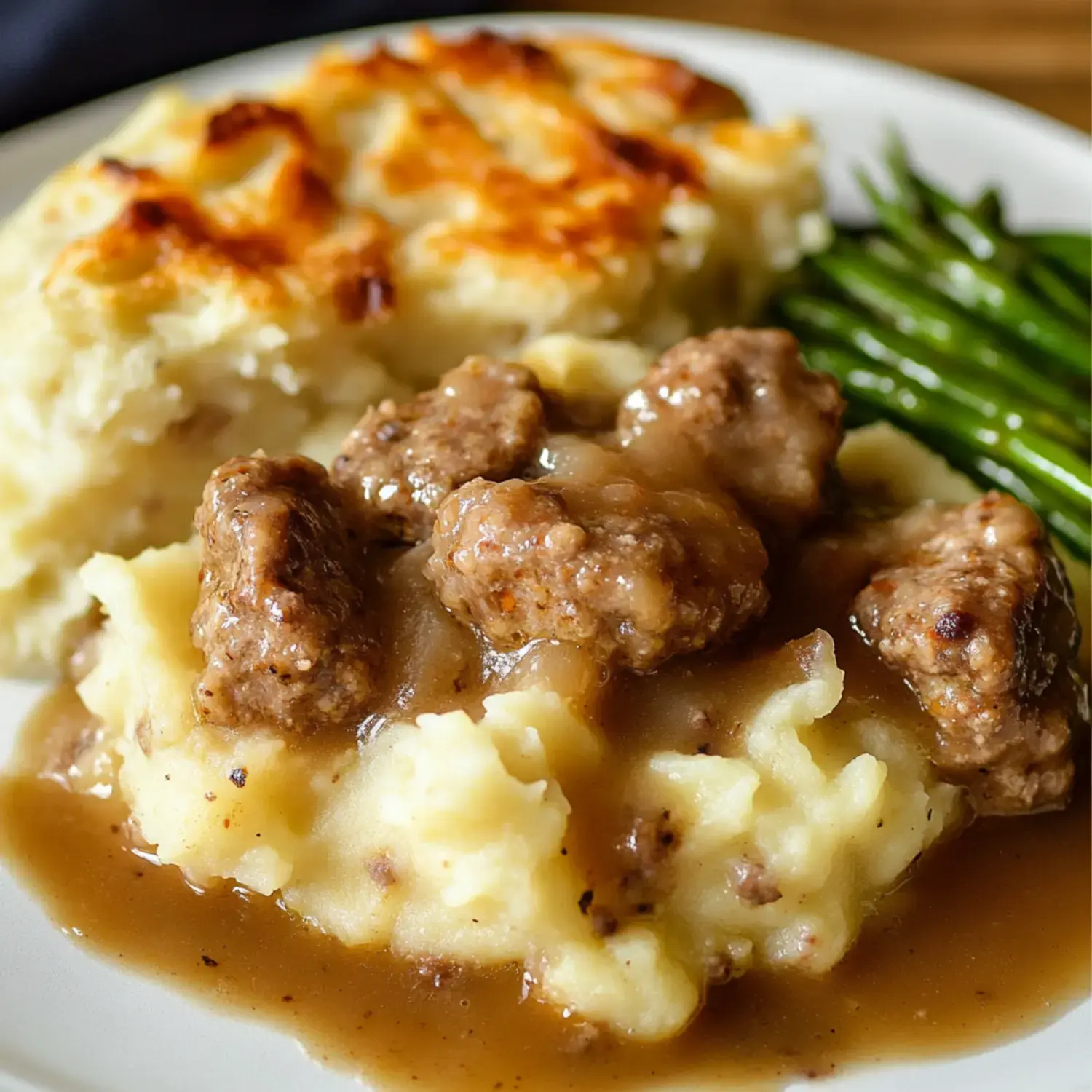 A plate of mashed potatoes topped with meatballs in gravy, accompanied by a side of green beans and a serving of cheesy baked potato.