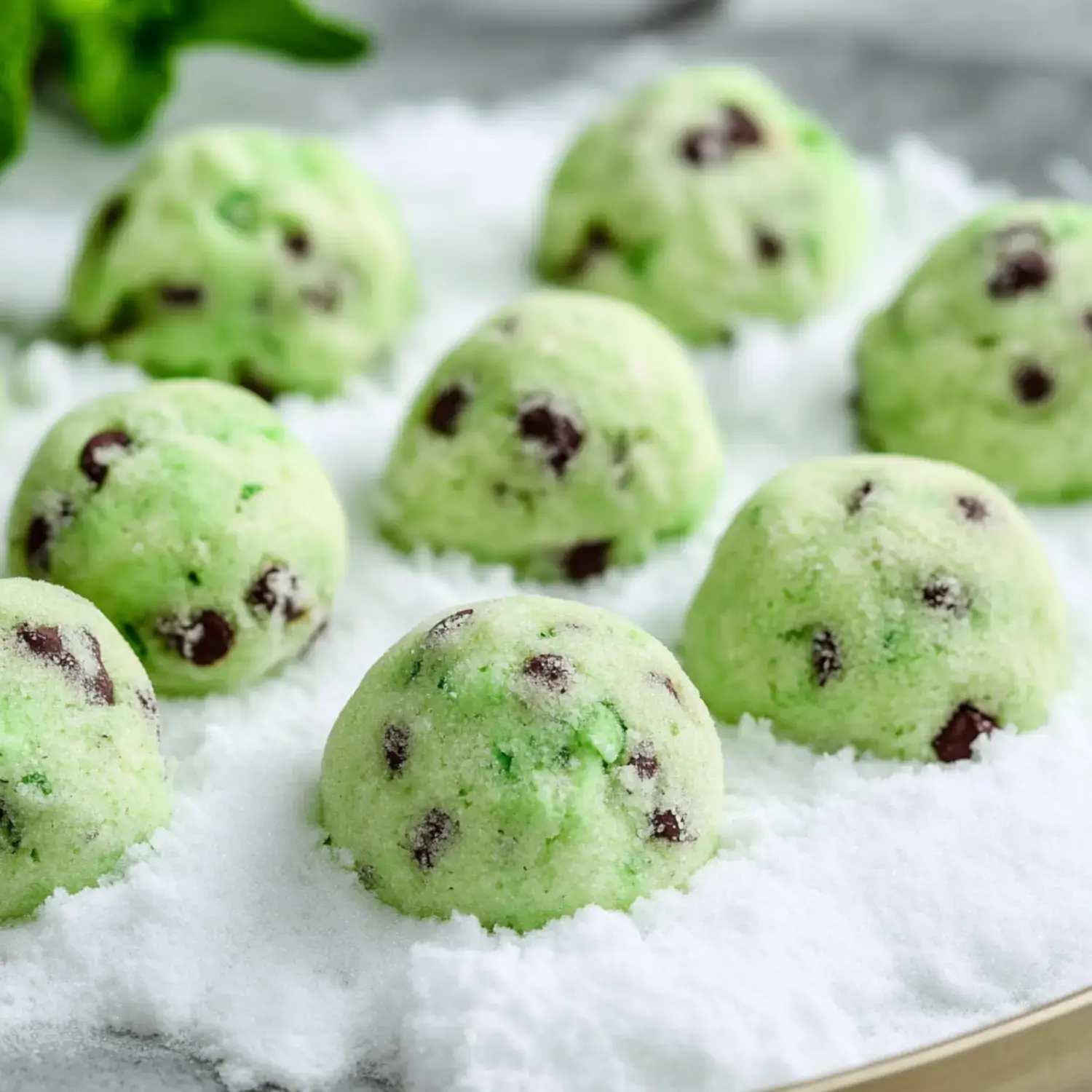 Eight green mint chocolate chip cookies are arranged on a bed of white sugar, resembling snow.