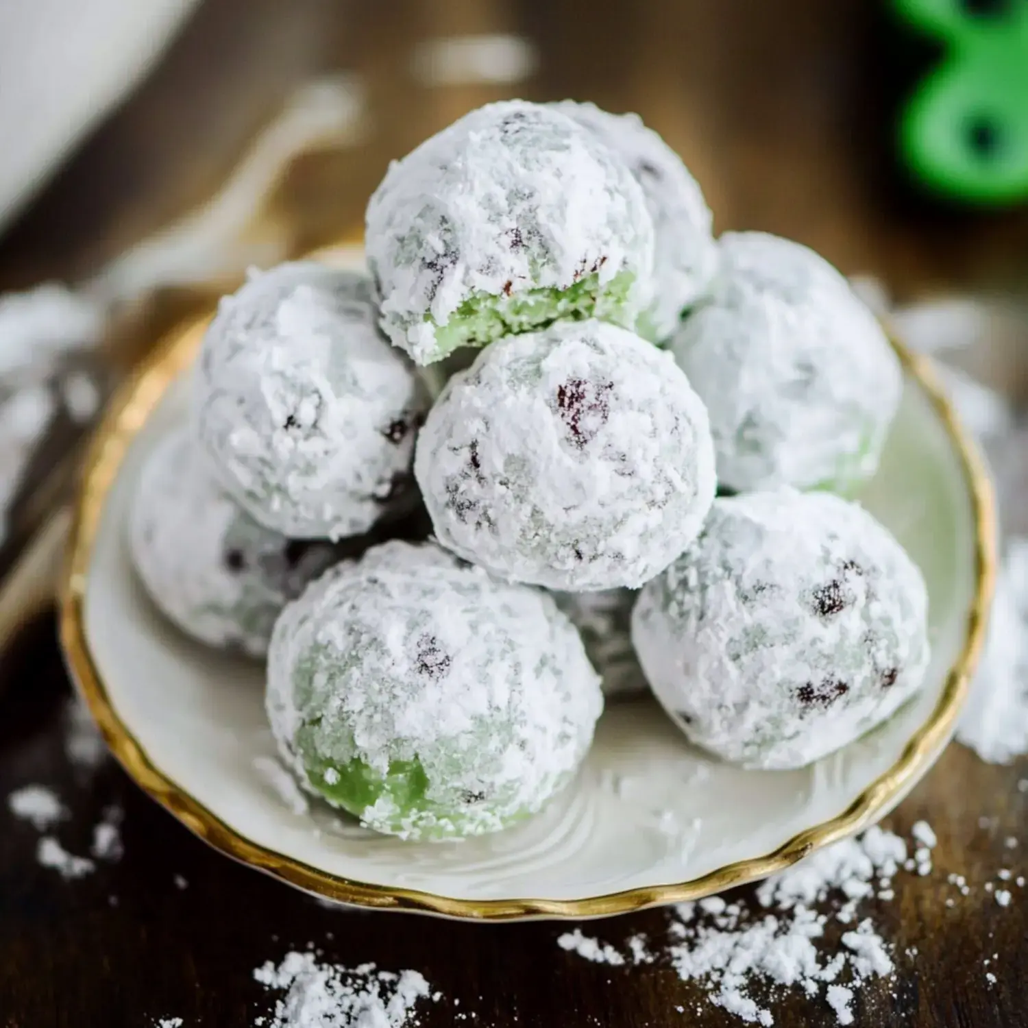 A plate of green and white powdered dessert balls, some with bites taken out, is displayed on a wooden surface with some powdered sugar scattered around.