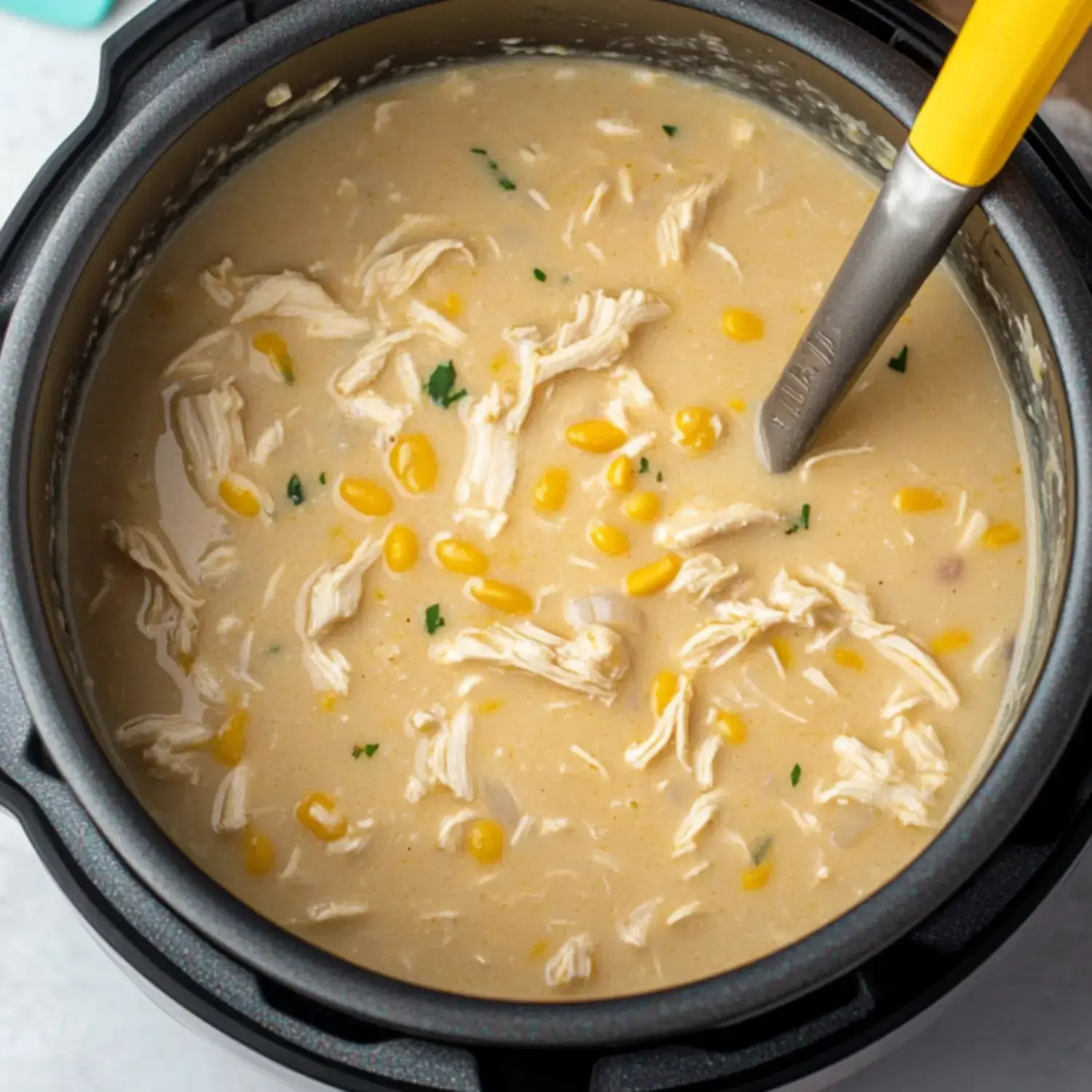 A creamy soup with shredded chicken and corn is cooking in a pot, accompanied by a silver spoon with a yellow handle.