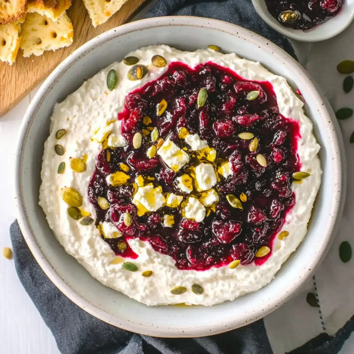 A bowl of whipped ricotta topped with a layer of berry compote, crumbled feta, pumpkin seeds, and a drizzle of olive oil, accompanied by pieces of toasted bread.