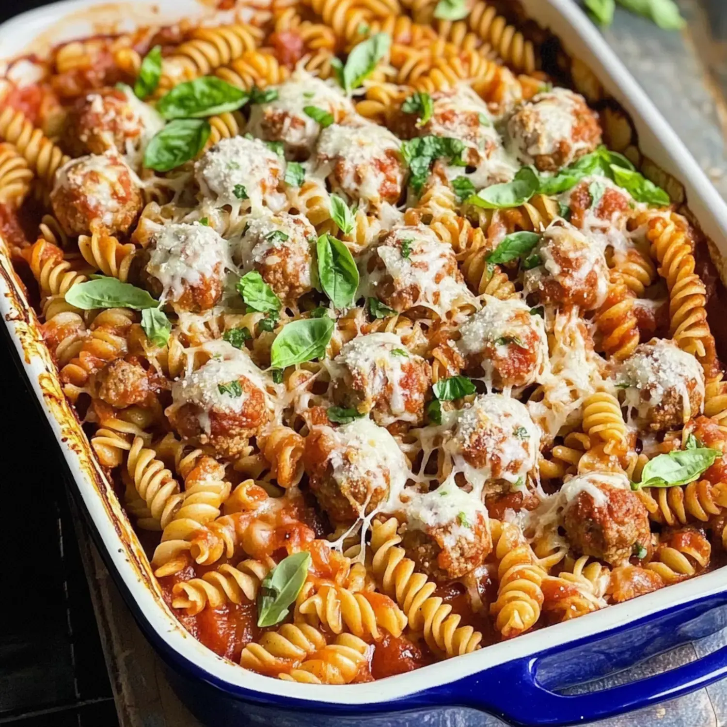 A baked dish of fusilli pasta topped with meatballs, marinara sauce, melted cheese, and fresh basil leaves.