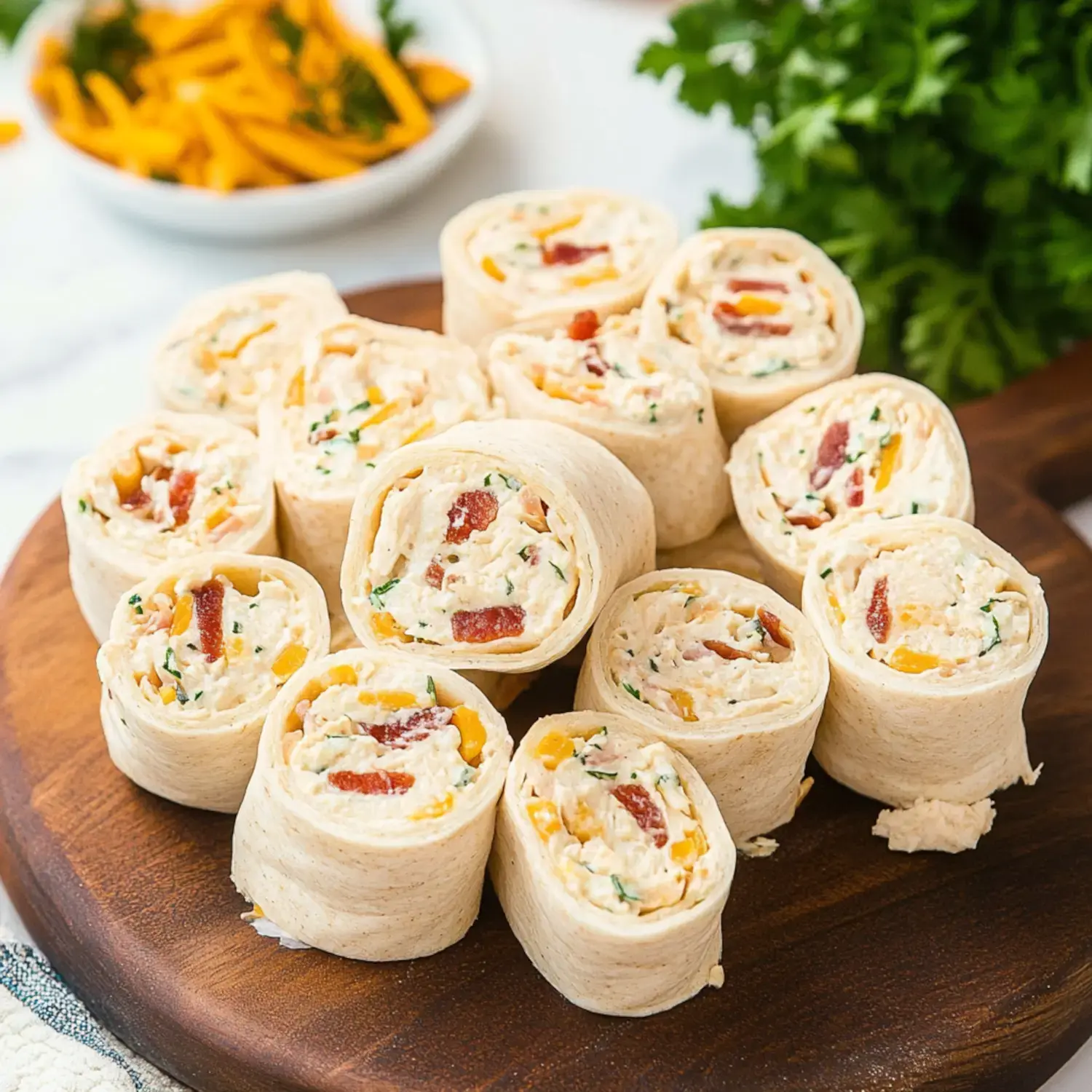 A wooden platter is filled with sliced tortilla pinwheels containing a creamy filling with bacon, herbs, and cheese, accompanied by a bowl of sliced peppers in the background.