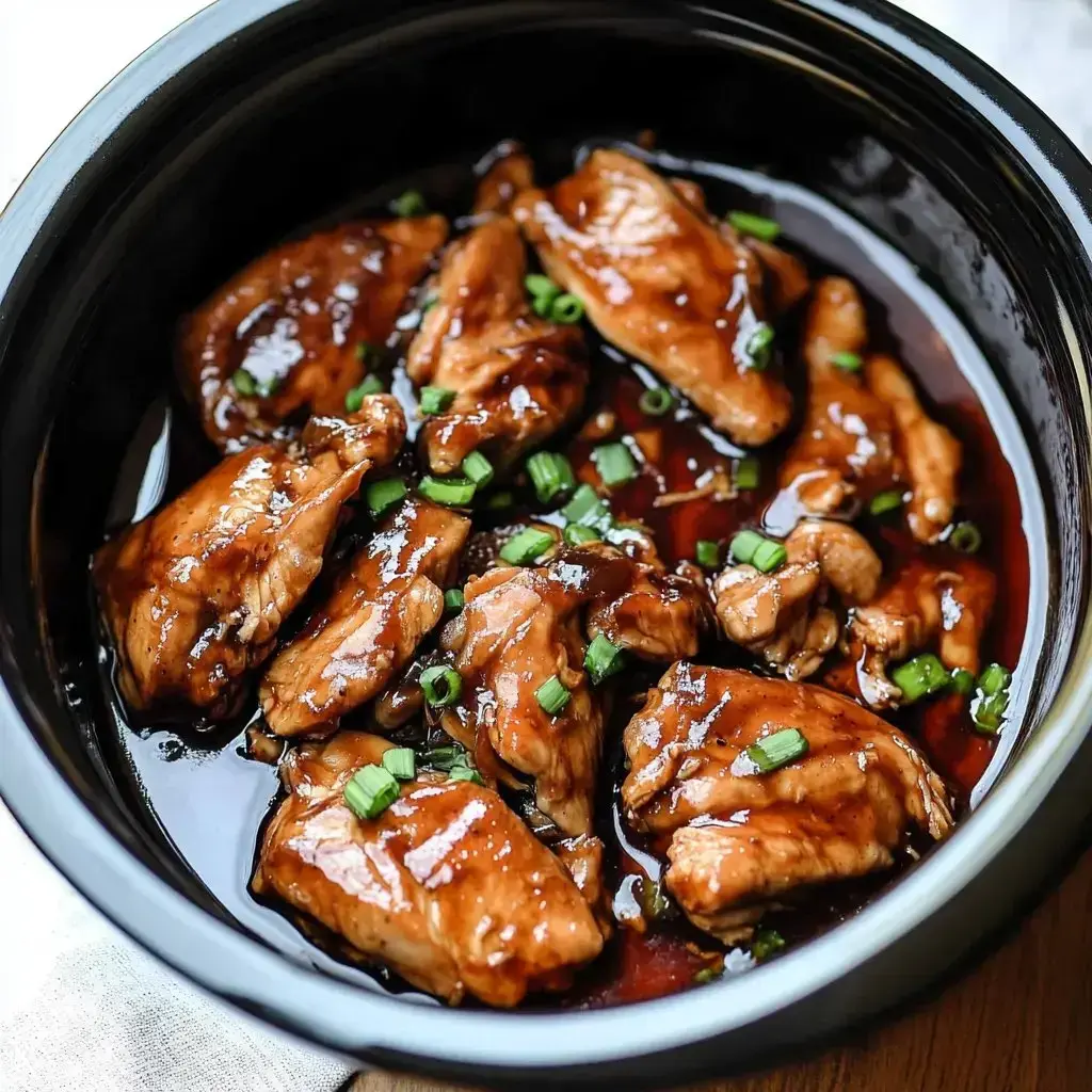 A close-up view of tender chicken pieces coated in a glossy sauce and garnished with chopped green onions in a black dish.