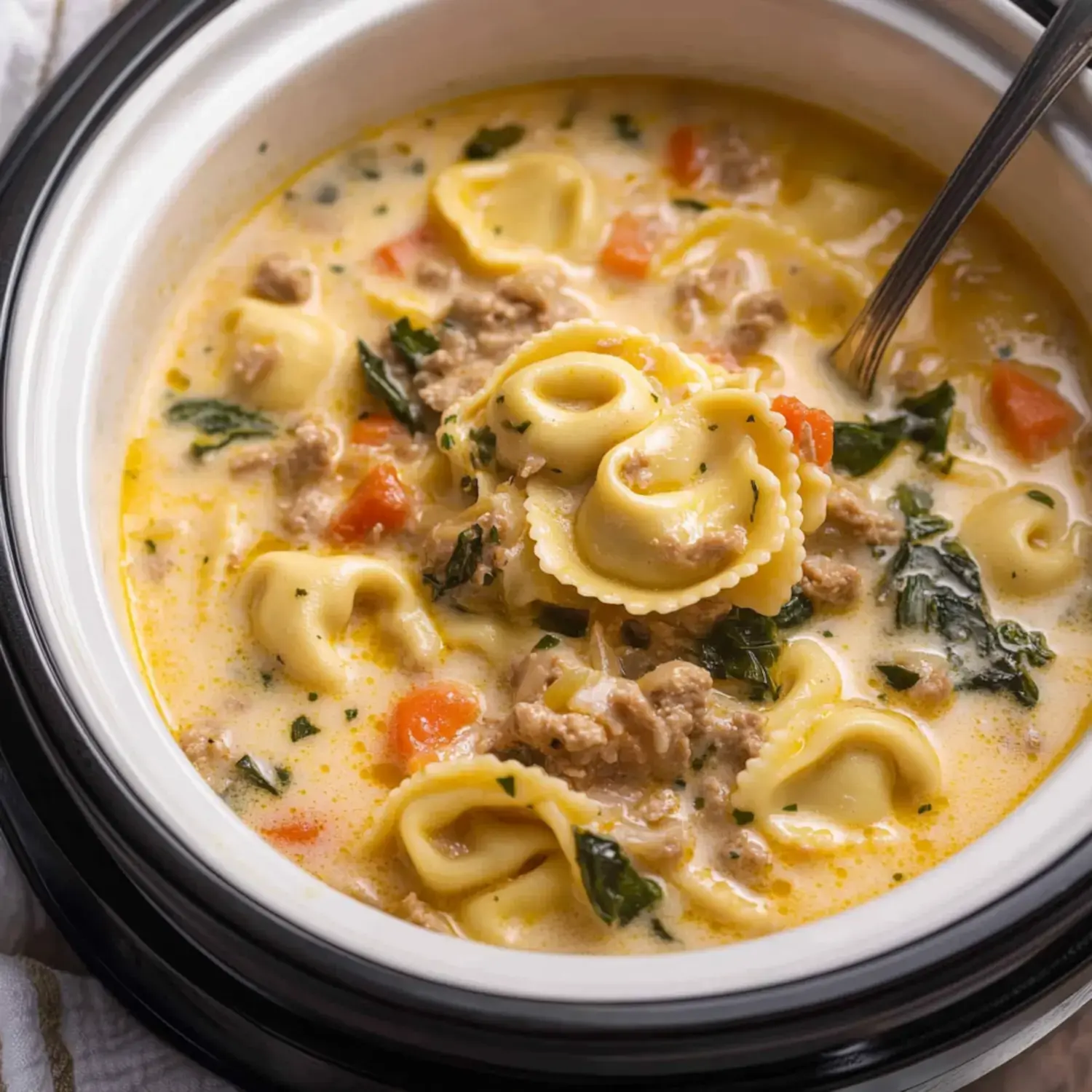 A creamy soup with pasta, ground meat, spinach, and diced vegetables is shown in a bowl with a spoon.