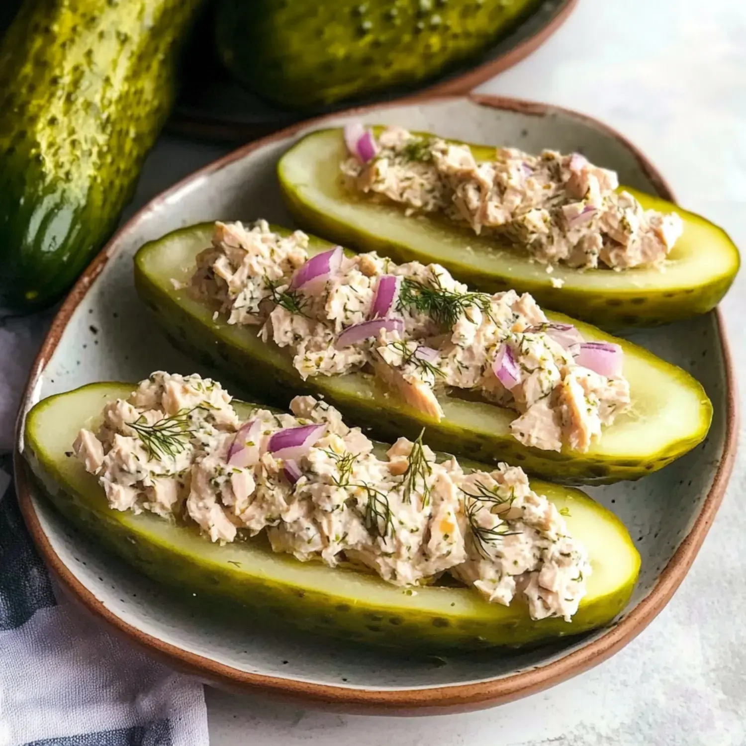 Three halved pickles filled with a creamy chicken salad garnished with red onion and fresh dill, served on a speckled plate.