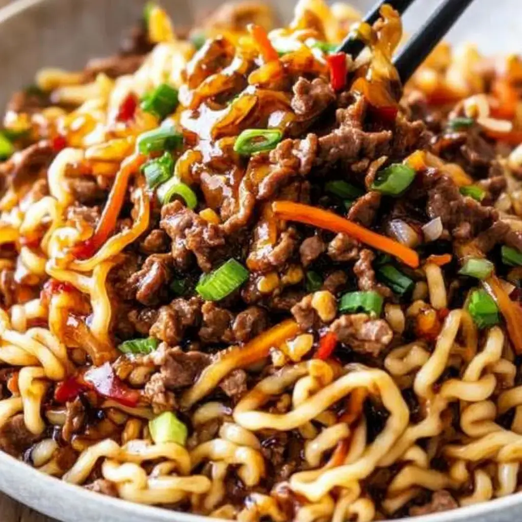 A close-up image of a bowl of noodles topped with beef, vegetables, and a glossy sauce, with chopsticks picking up a portion.