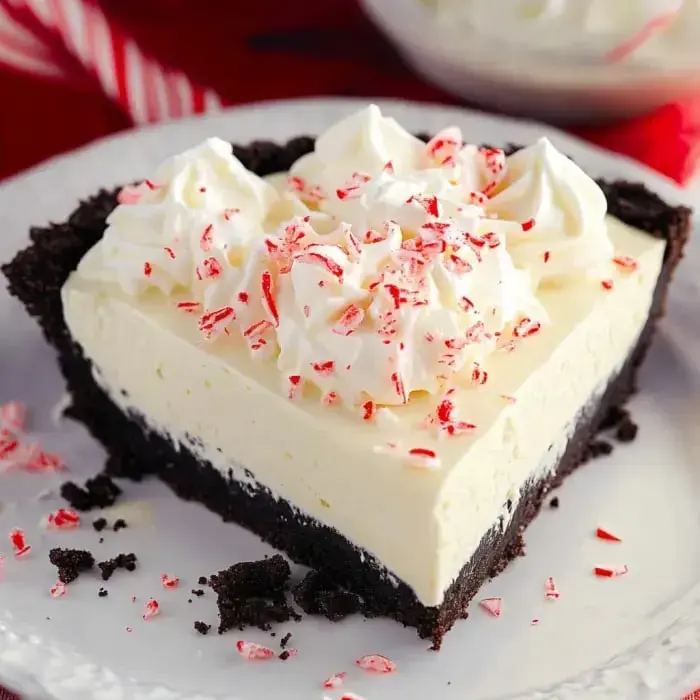 A slice of peppermint cream pie topped with whipped cream and crushed peppermint candies on a decorative plate.