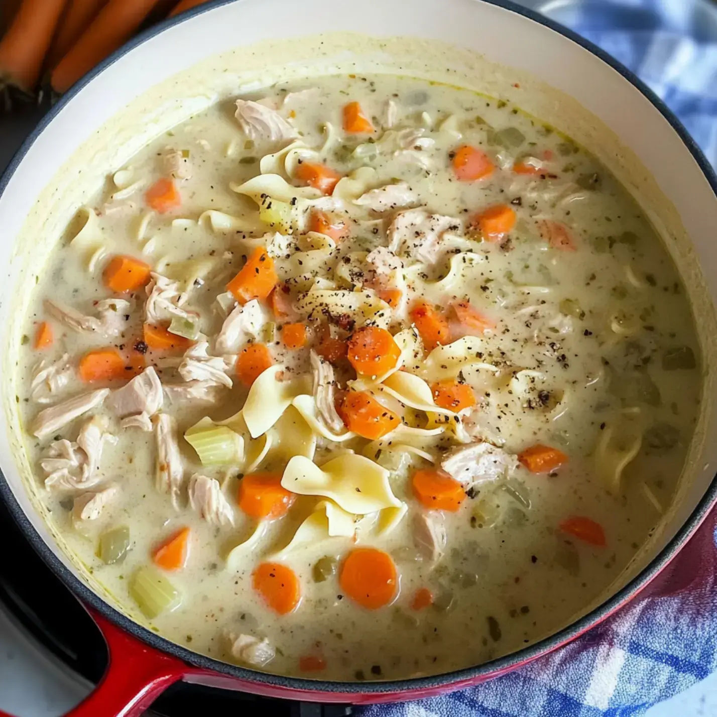 A close-up view of a pot filled with creamy chicken noodle soup, featuring pieces of chicken, carrots, and egg noodles topped with black pepper.