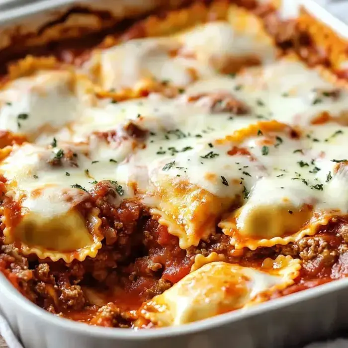 A close-up of a baked dish featuring layers of meat, cheese, and ravioli topped with melted mozzarella and herbs.