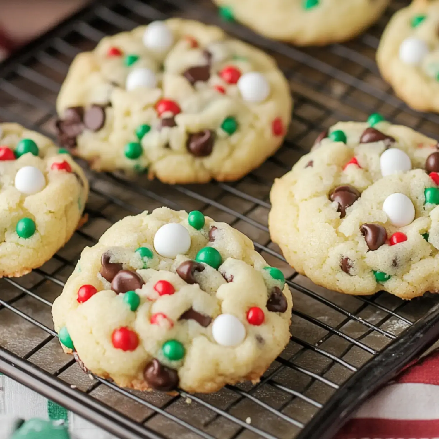 A close-up of colorful holiday cookies with chocolate chips and festive red, green, and white decorations, arranged on a wire rack.