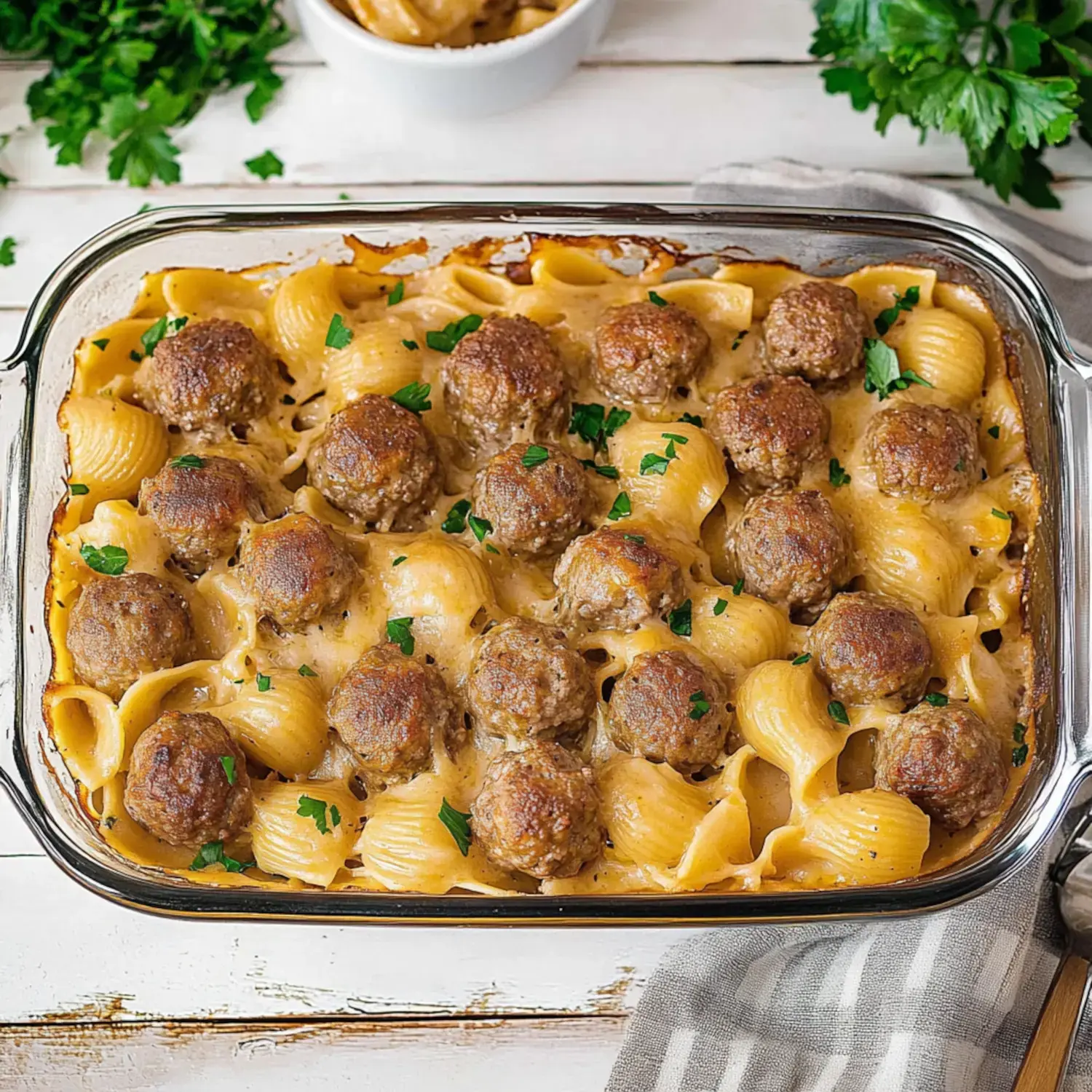 A baked casserole dish filled with pasta shells and meatballs, topped with a creamy sauce and garnished with parsley.