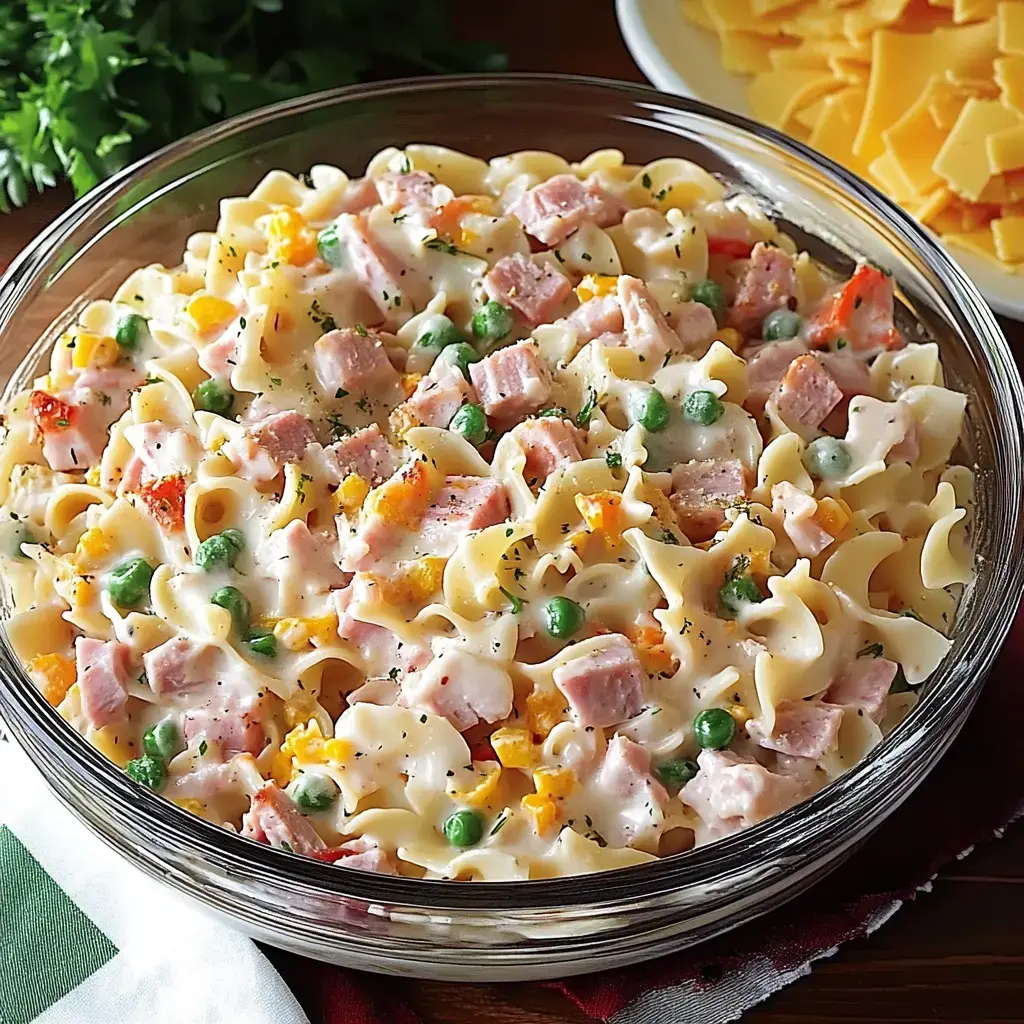 A clear glass dish filled with creamy pasta mixed with ham, peas, and cheese.