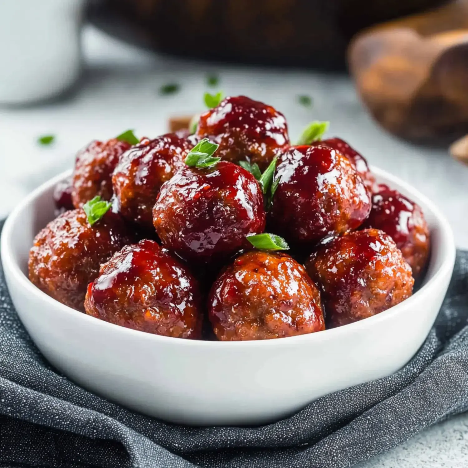 A close-up image of a bowl filled with glossy meatballs, garnished with green herbs.