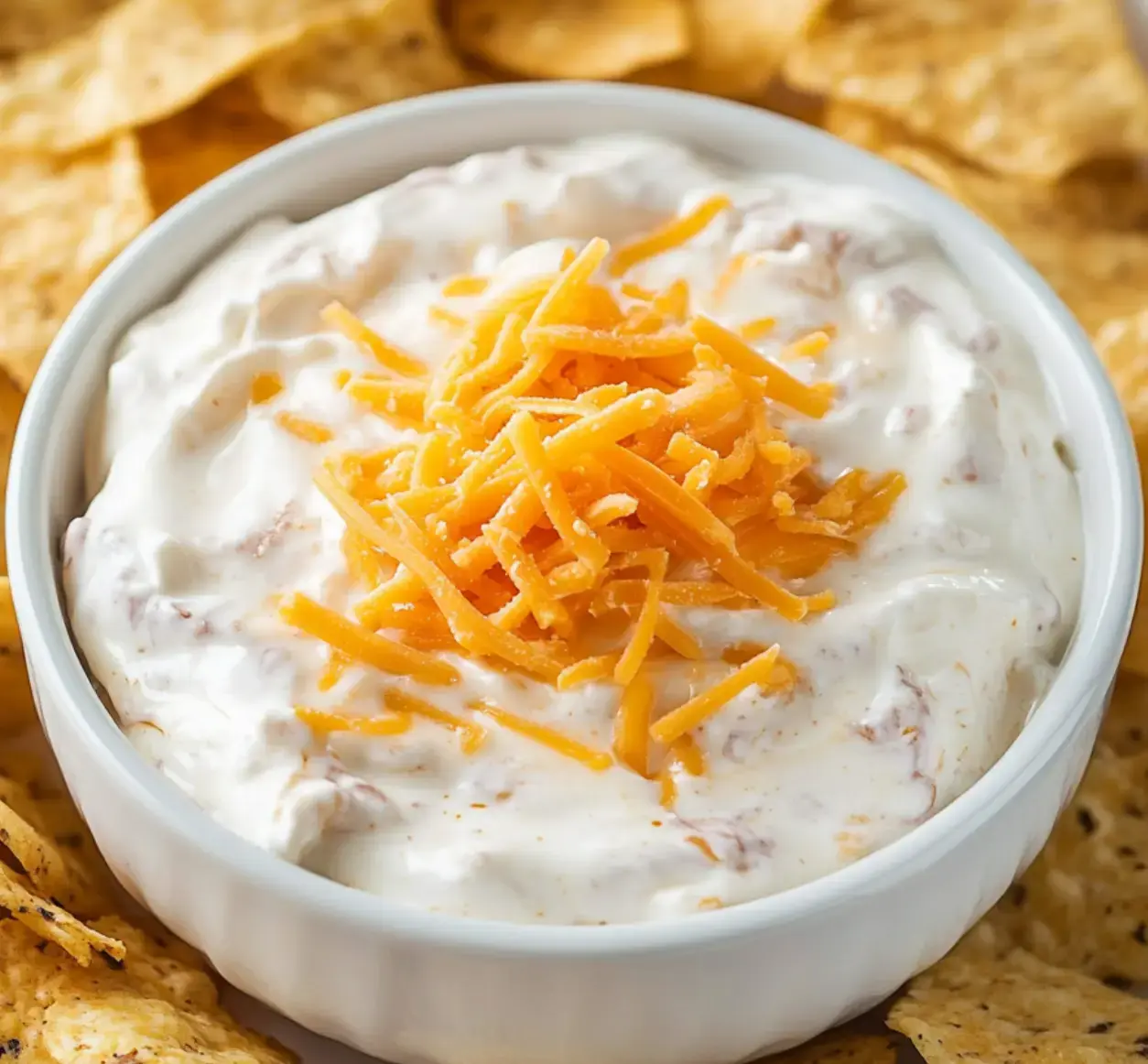 A white bowl filled with creamy dip, topped with shredded cheddar cheese, surrounded by crispy tortilla chips.