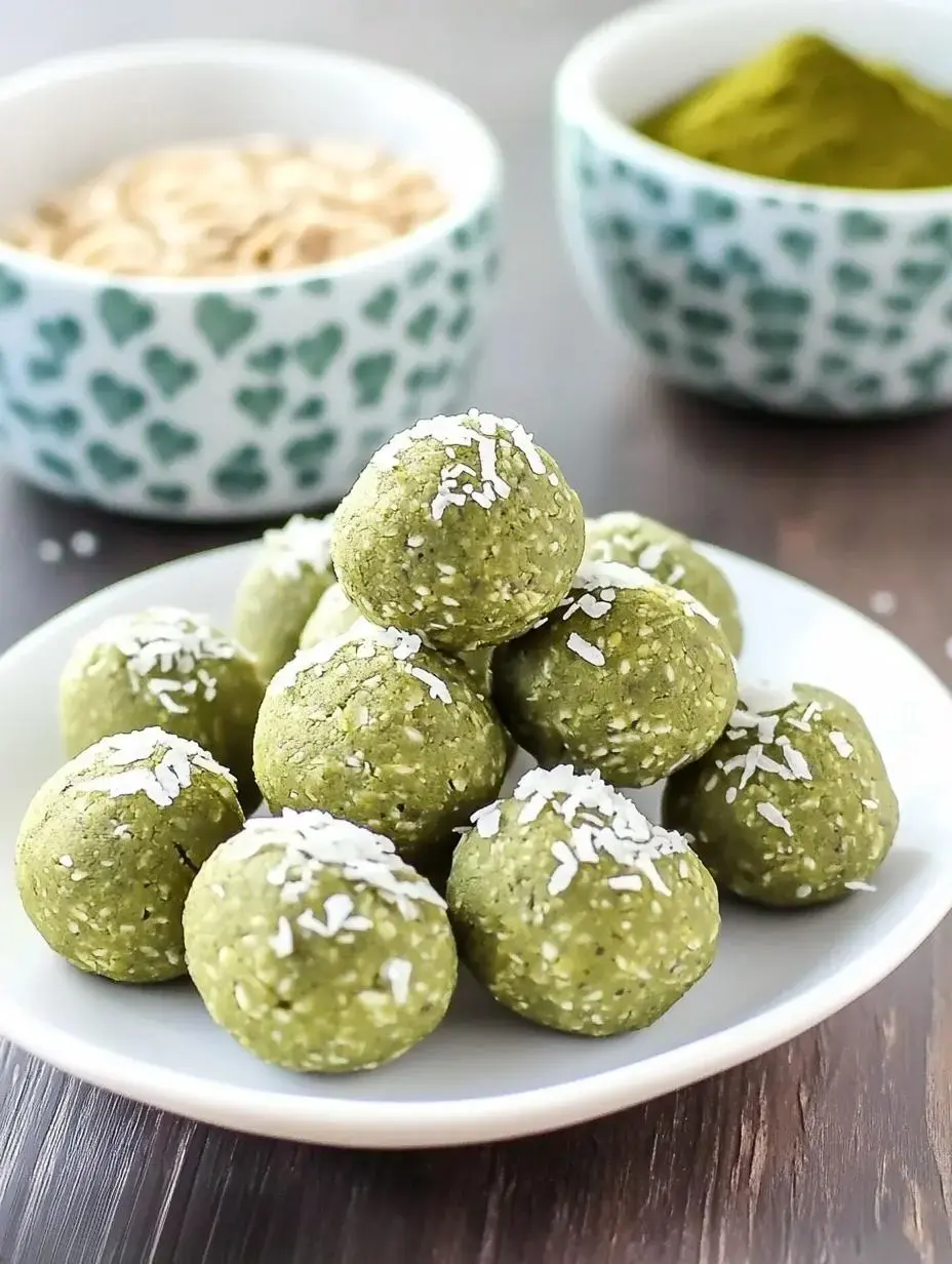 A plate of green energy balls topped with shredded coconut, with two bowls of matcha powder and oats in the background.