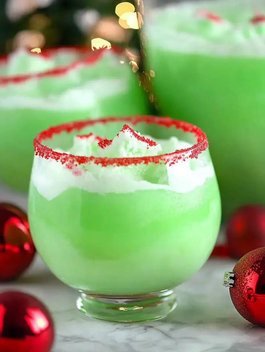 A festive green drink with whipped topping and red sugar rim is placed beside shiny red ornaments on a marble surface.