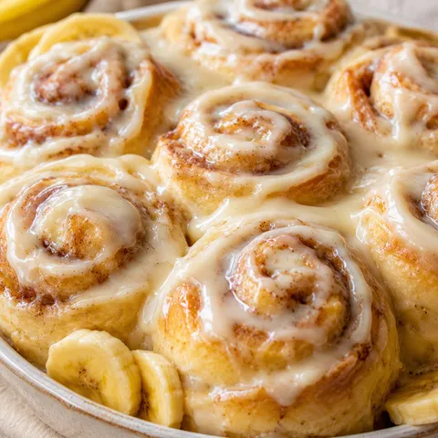 Banana cinnamon rolls with cream cheese icing on a light wood board