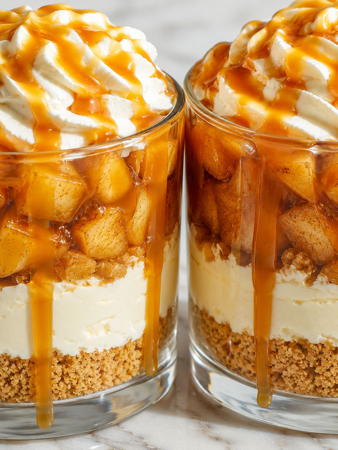 Two caramel apple dessert cups with cream cheese layers, spiced apples, and caramel drizzle