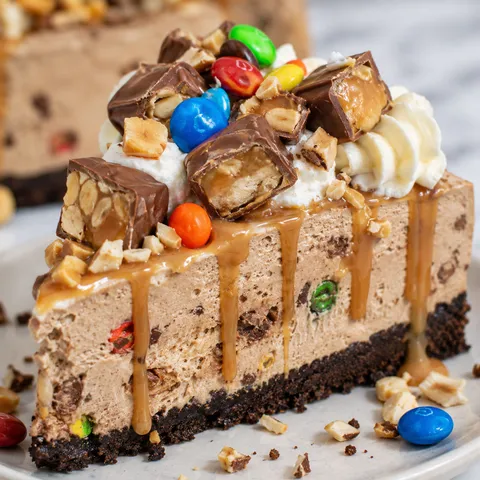 M&M Snickers cheesecake slice with caramel, whipped cream, and peanuts
