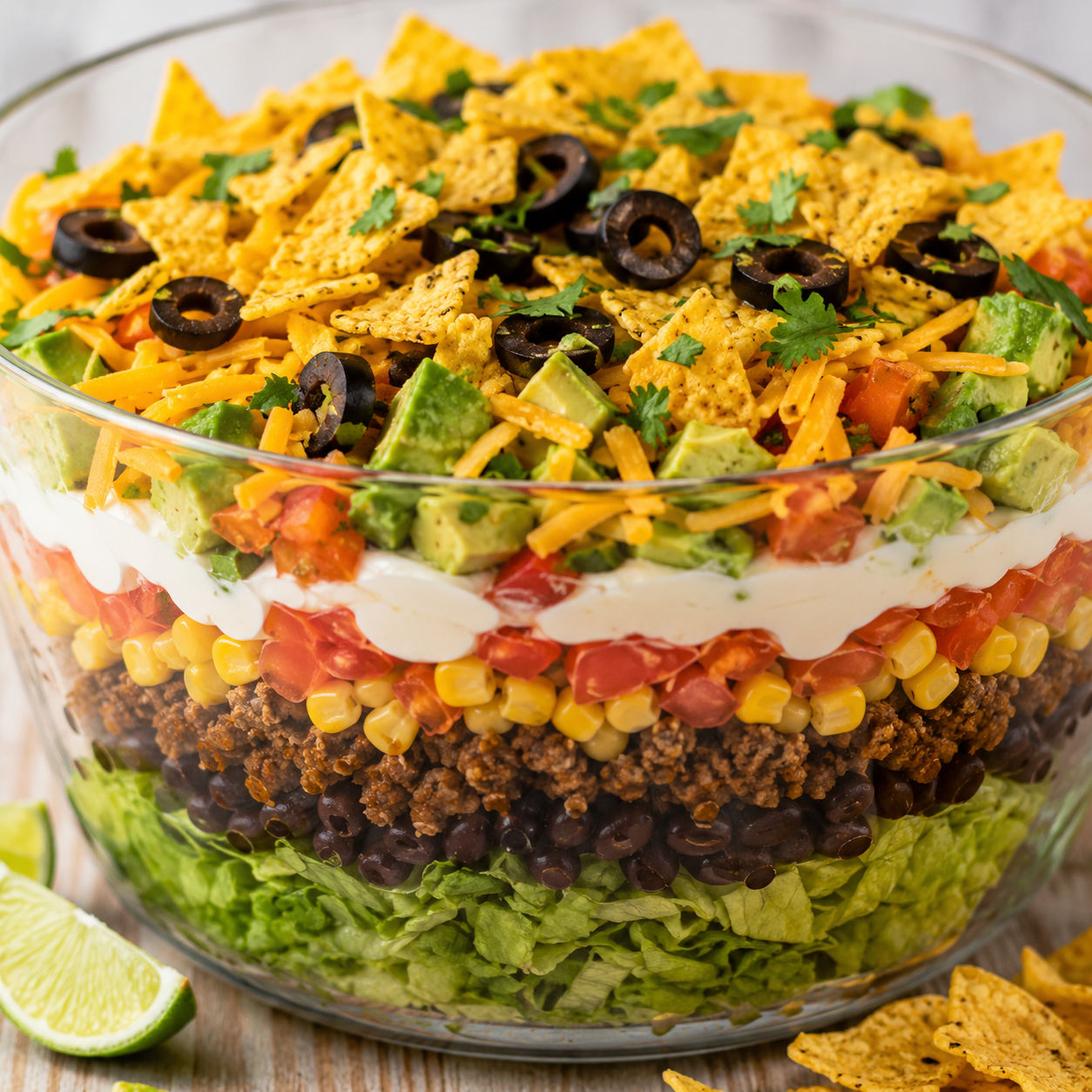 Seven-Layer Taco Salad in a glass bowl with avocado, olives, and chips
