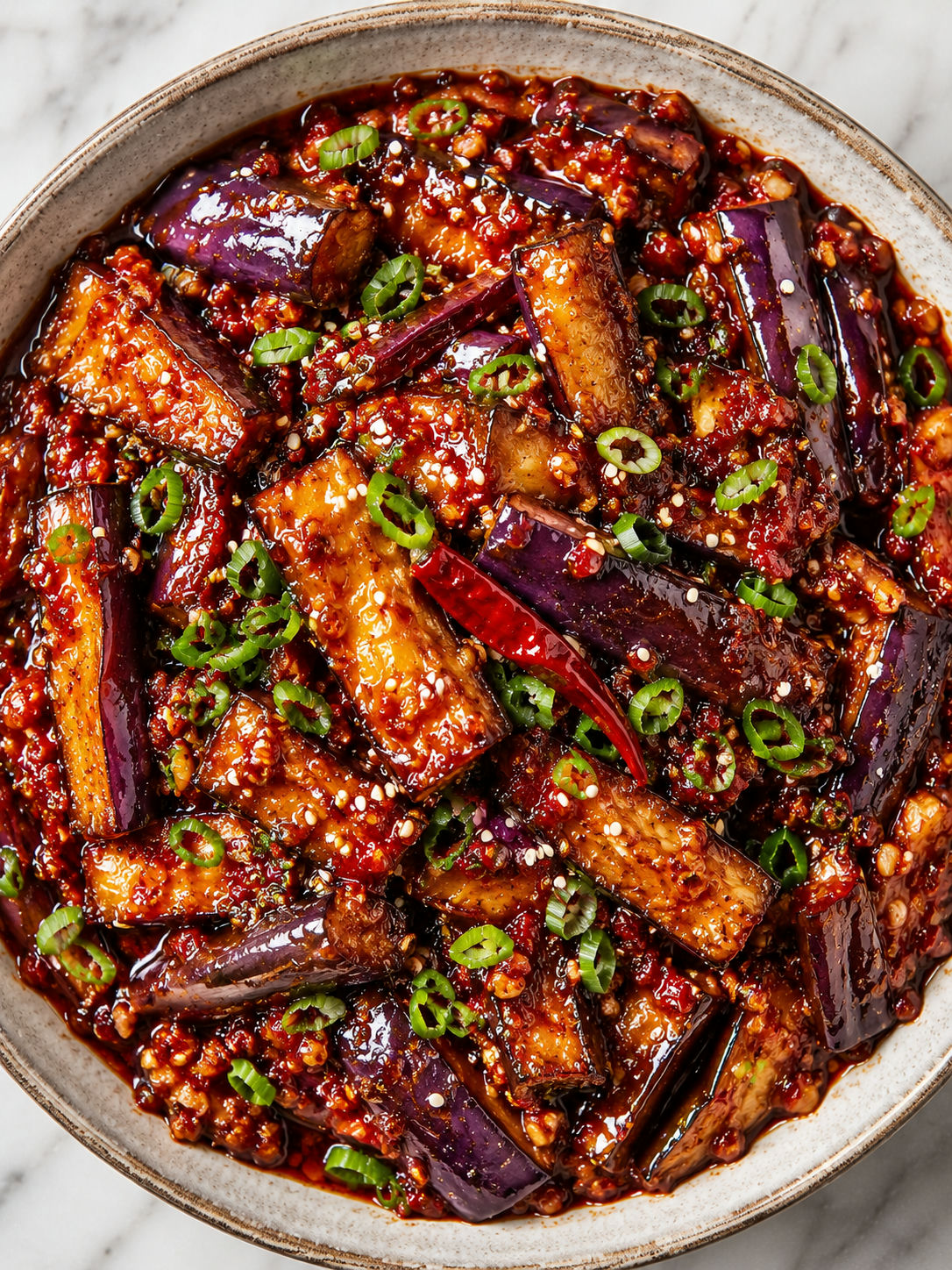 Top-down bowl of spicy Sichuan eggplant with glossy chili sauce, scallions, and sesame seeds