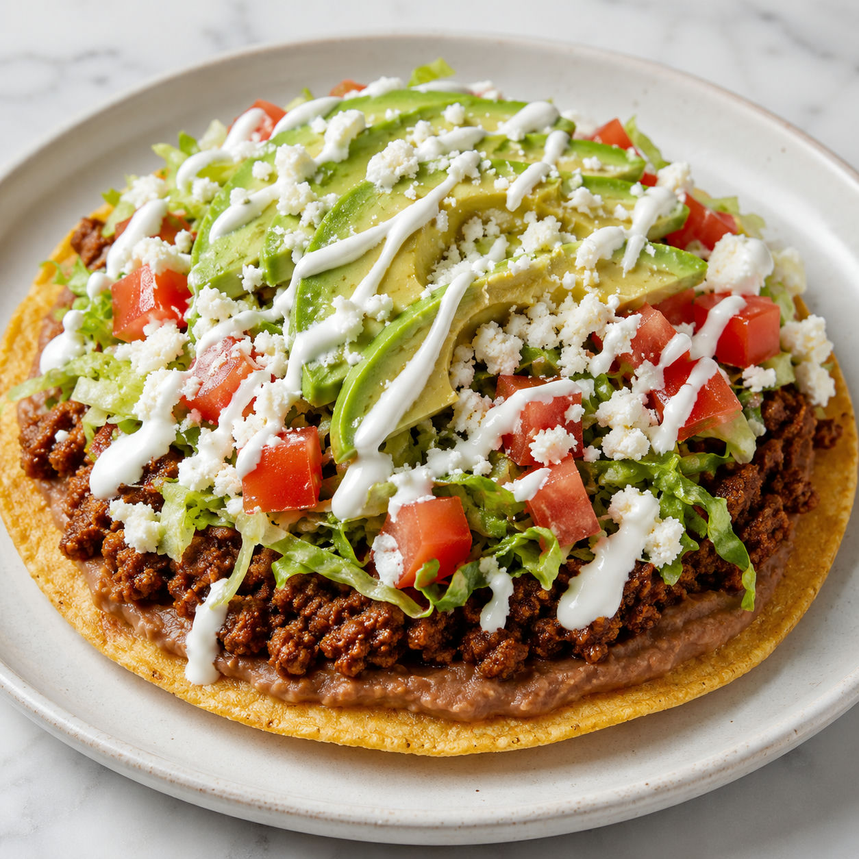 Ground beef tostada loaded with refried beans, avocado, queso fresco, and crema on a ceramic plate