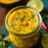 A jar of yellow sauce with limes and chili peppers on the side.