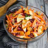 A bowl of vegetables, including carrots and apples, sits on a table.