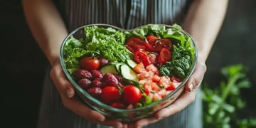 A person is holding a heart-shaped bowl filled with a colorful variety of fresh vegetables and greens.