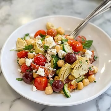 A vibrant salad featuring cherry tomatoes, cucumber, chickpeas, olives, artichokes, feta cheese, and fresh herbs, served in a white bowl.