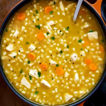 A close-up view of a bowl of soup featuring chicken, carrots, and small grains in a yellow broth, garnished with herbs and black pepper.