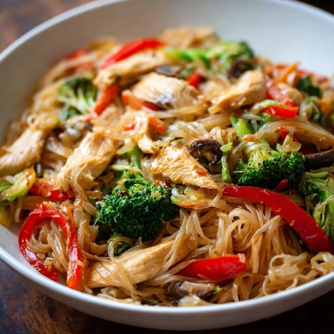 A bowl of chicken stir fry with rice noodles, broccoli, and peppers.